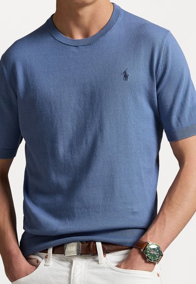 Pull court en tricot bleu clair à manches courtes avec un col rond et un petit logo brodé sur la poitrine. Porté avec un pantalon blanc et une ceinture marron.