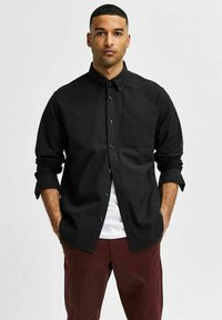 Camisa negra de botones con cuello, de manga larga arremangada en los puños, y un bolsillo en el pecho, llevada sobre una camiseta blanca y pantalones color burdeos.