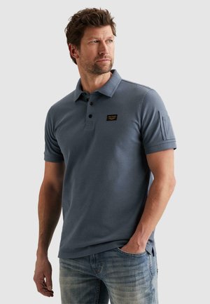 Mann mit Bart und zerzaustem Haar, der ein kurzärmeliges Poloshirt in Schieferblau mit Logo-Patch auf der Brust und Tasche am linken Ärmel trägt, kombiniert mit ausgewaschenen Jeans.