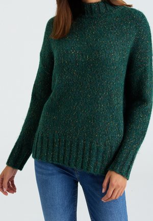 Vrouw draagt een donkergroene gebreide trui met spikkels, geribbelde manchetten en zoom, gecombineerd met een blauwe spijkerbroek, tegen een effen achtergrond.