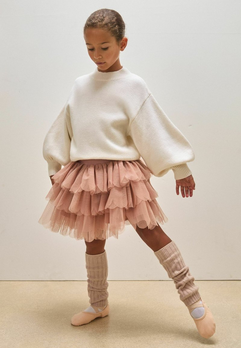 Maglione bianco con maniche a palloncino, gonna in tulle rosa a strati, calze a coste fino al ginocchio e scarpette da balletto rosa. Sfondo neutro.
