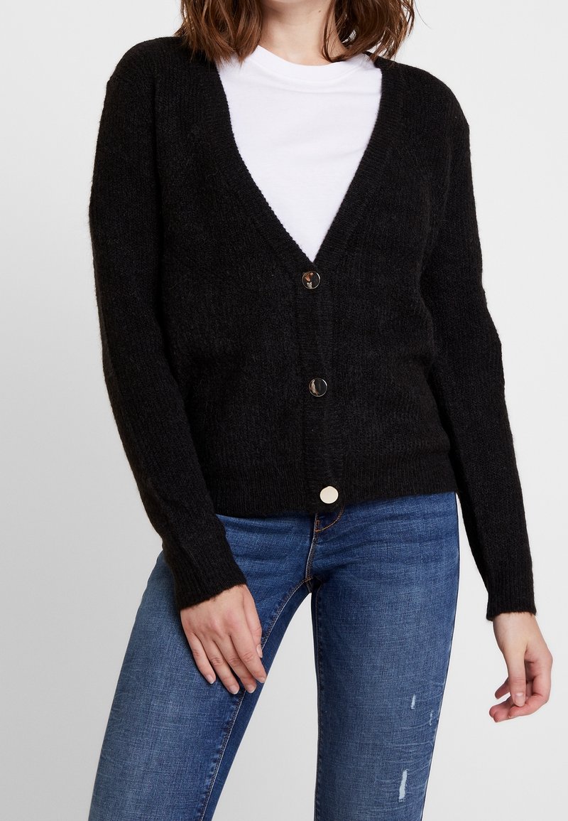 Woman wearing a black buttoned cardigan over a white shirt with blue skinny jeans, standing with hands near hips against white background.