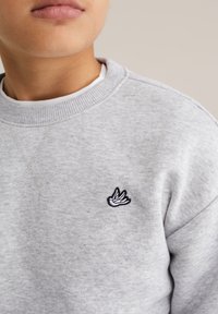 Szürke pulóver kerek nyakkivágással és bordázott szegélyekkel. A bal mellrészen egy kis fekete-fehér hímzett madár logó található.