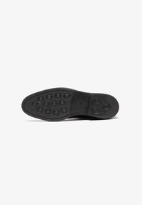 Suela de zapato de goma negra con agarres circulares y bordes texturizados. Presenta una sección central lisa y un diseño de punta redondeada.