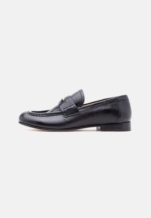 Mocasín de cuero negro para hombre con tacón bajo y una correa decorativa sobre el empeine, mostrado en perfil lateral sobre un fondo liso.