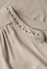 Béžová látka s vertikálním žebrováním má nabíraný detail a dekorativní stříbrné nýty kolem zaobleného výstřihu. Hladká textura a jemný lesk.