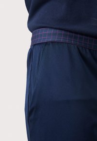 Ναυτικές βερμούδες με λείο υλικό, ελαστική μέση με μοτίβο μωβ πλέγματος και πλευρικές τσέπες για πρακτικότητα.