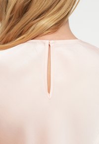 Απαλό ροζ μπλούζα με άνοιγμα σε σχήμα κλειδιού στην πλάτη, με λεπτή ραφή κατά μήκος του ντεκολτέ και απαλό, λείο ύφασμα.