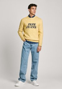 Rumena pletena majica s logotipom "PEPE JEANS", v kombinaciji z svetlo modrimi kavbojkami in belimi športnimi čevlji. Majica ima okroglo izrezano ovratnik in rebraste manšete.