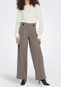 Breda byxor i strukturerad beige tyg, med dubbla framknappar, kombinerat med en vit stickad tröja. Svarta klackskor fulländar outfiten.