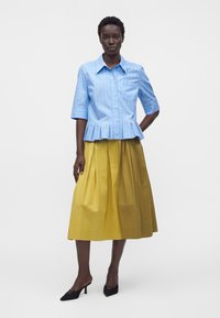 Femme portant une chemise bleue à manches courtes boutonnée avec un ourlet plissé et une jupe midi plissée jaune, debout devant un fond uni.
