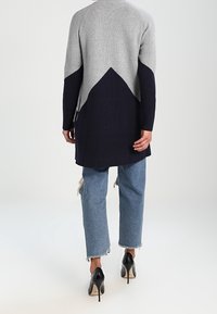 Langer gestrickter Cardigan mit grauem Oberteil und dunkelblauem Unterteil, verziert mit einem geometrischen Muster. Kombiniert mit zerrissenen Jeans und High Heels.