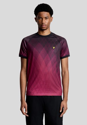 Giovane uomo che guarda avanti indossando una maglietta a maniche corte con motivo argyle bordeaux e nero e un piccolo logo di un uccellino giallo sul petto.