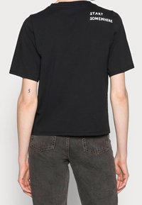 Camiseta de algodón negra con mangas cortas, con un cuello redondo y texto blanco en la parte de atrás que dice "EMPIEZA EN ALGÚN LUGAR". Diseño simple y sin adornos.