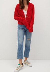 Cardigan rouge en maille torsadée avec un col en V et deux boutons dorés, associé à un jean bleu clair déchiré et court, ainsi qu'à des baskets multicolores.