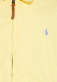 Gelbe Zip-Jacke aus weichem Material mit einem blauen gestickten Logo eines Pferdes und Reiters auf der linken Brust. Reißverschluss mit braunem Lederanhänger.