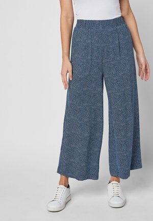 Frau trägt blaue, weit geschnittene Hosen mit weißen abstrakten Punkte-Mustern und weißen Sneakers, steht vor einem schlichten grauen Hintergrund.