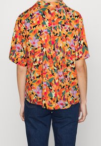 Frau mit einer kurzärmeligen orangefarbenen Bluse mit einem bunten abstrakten Blumenmuster und dunkelblauen Jeans, von hinten gezeigt.