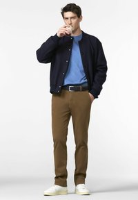 Young man wearing a navy jacket, blue shirt, brown pants, and white sneakers drinking from a small glass cup.
