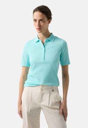 Kobieta w jasnoniebieskiej koszulce polo z krótkim rękawem i guzikami, połączona z beżowymi spodniami z wysokim stanem na jednolitym tle.