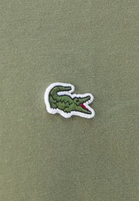 Zelená bavlnená látka obsahuje vyšívané logo krokodíla s bielym okrajom a červenými akcentmi, umiestnené na ľavej strane.