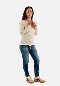 Haut à capuche beige à manches longues avec un cordon de serrage à la taille, assorti à un jean skinny bleu et des baskets beiges avec des rayures bordeaux.
