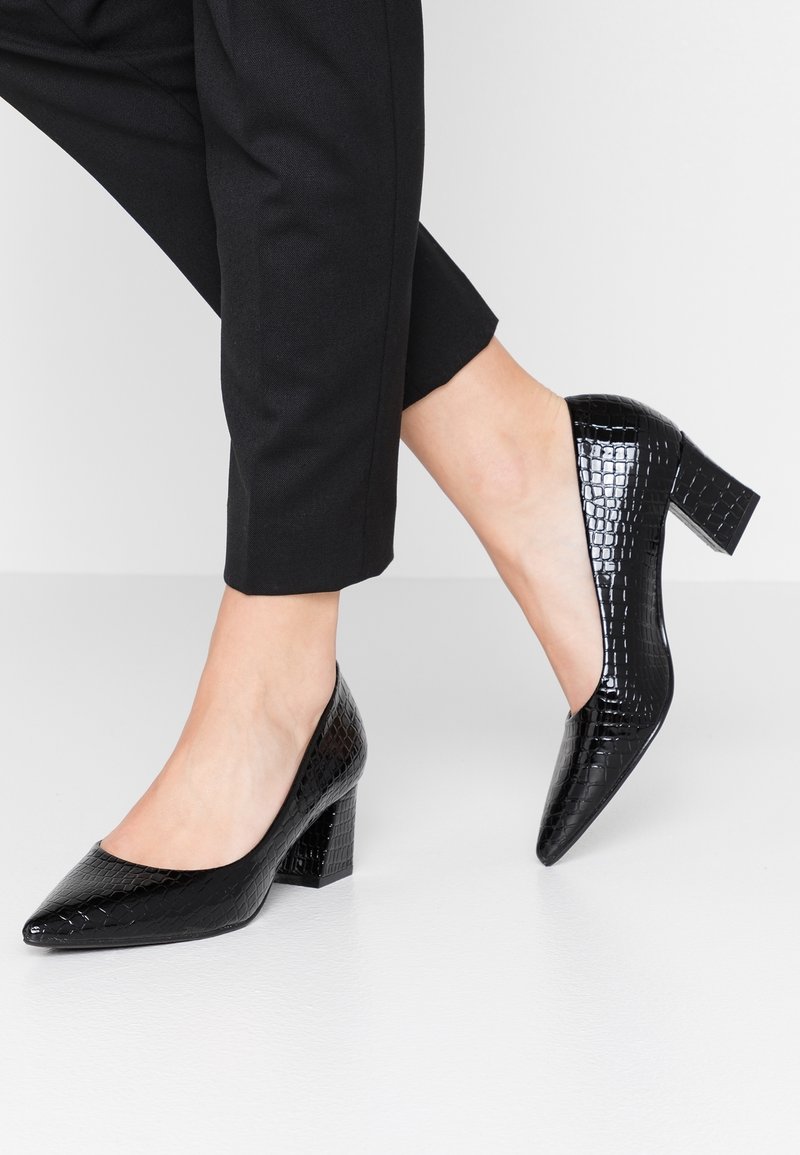 Chaussures en cuir verni noir à talon moyen avec texture crocodile portées avec un pantalon noir coupé au-dessus de la cheville, un pied à plat et l’autre légèrement relevé.