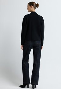 Kobieta z kokiem, ubrana w czarny sweter z długim rękawem, ciemnoniebieskie dżinsy prostego kroju i czarne botki na obcasie, stojąca na białym tle.