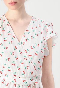 White wrap dress with a cherry pattern in red and green, featuring a V-neckline and flutter sleeves. Textured fabric with a flowy fit.