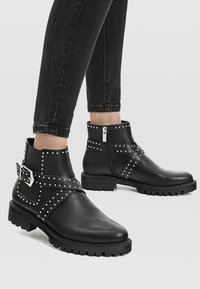 Bottes noires jusqu'à la cheville en cuir lisse, ornées de clous en argent et d'une large sangle avec une boucle. Semelle en caoutchouc crantée pour une meilleure adhérence.