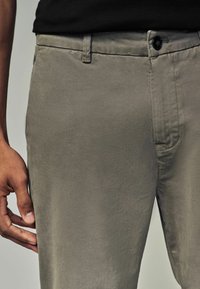 Graue Baumwollchinos mit einer glatten Textur, ausgestattet mit einem Knopfverschluss und seitlichen Taschen, teilweise gezeigt neben einer Hand, die auf dem Stoff ruht.