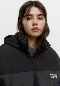 Giacca nera imbottita con cappuccio; presenta una chiusura con zip, una texture liscia e un piccolo logo sul lato sinistro.