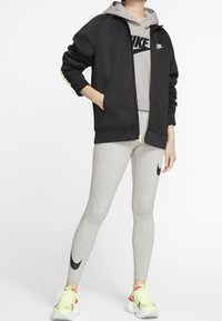 Černá zipová bunda s zelenými akcenty, šedá mikina pod ní a světle šedé legíny se černým logem. Oblečení doplňují jasné boty.