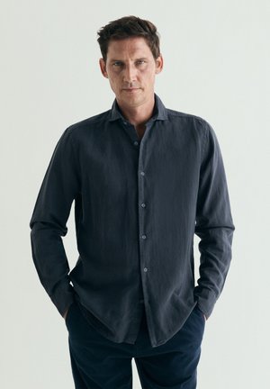Hombre con el pelo corto y castaño, vestido con una camisa de botones gris oscuro y pantalones oscuros, de pie con las manos en los bolsillos frente a un fondo claro y sencillo.