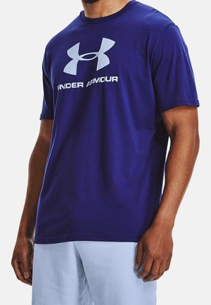 Mężczyzna ubrany w ciemnoniebieską koszulkę Under Armour z logo i napisem, w zestawieniu z jasnoniebieskimi spodenkami, na jednolitym tle.