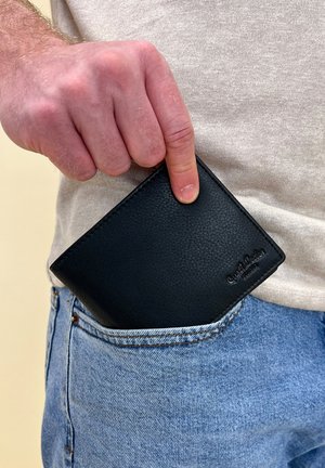 Black leather wallet partly in a jeans pocket, showcasing pebbled texture and a smooth edge. Logo embossed in the bottom right corner.