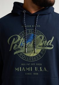 Námořnická modrá mikina s kapucí vyrobená z bavlny s měkkou texturou. Má zelený grafický text, šňůrky s stříbrnou kovovou součástí a přední kapsu.