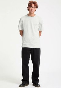 T-shirt cinzento claro de manga curta com um pequeno logótipo no peito, combinada com calças pretas largas e sapatos pretos.