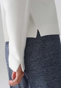 White knit sweater with side slits, paired with textured navy pants featuring a diagonal pattern. Hand resting on the pants.