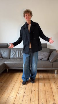 Casaco preto estilo peacoat com abotoamento duplo, camisa bege por baixo, jeans azuis largos e sapatos pretos. Em pé numa sala de estar com chão de madeira.