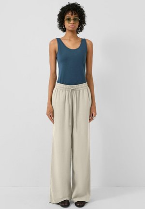 Femme aux cheveux bouclés portant des lunettes de soleil teintées vert, un haut sans manches bleu, un pantalon beige à jambes larges et des chaussures noires, debout devant un fond gris.