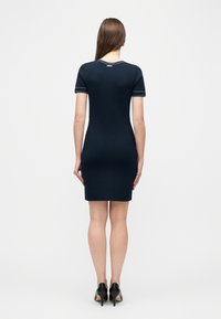 Γυναίκα με μακριά καστανά μαλλιά που στέκεται με την πλάτη προς τα εμπρός, φοράει εφαρμοστό κοντομάνικο φόρεμα σε ναυτικό μπλε και μαύρα ψηλοτάκουνα παπούτσια σε λευκό φόντο.