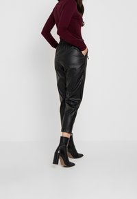 Zwarte leren joggingsbroeken met een elastische tailleband en zijzakken, gecombineerd met zwarte haklaarzen, gezien vanaf de achterkant.