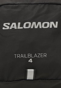 Fekete hátizsák, amelyen fehér színben a "SALOMON" márkanevet és a "TRAILBLAZER 4" feliratot láthatjuk, tartós anyagból készült, elegáns, matt felülettel.