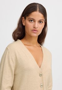 Cardigan beige avec un décolleté en V profond, des manches longues et trois grands boutons brillants sur le devant. Texture douce, matériau léger.