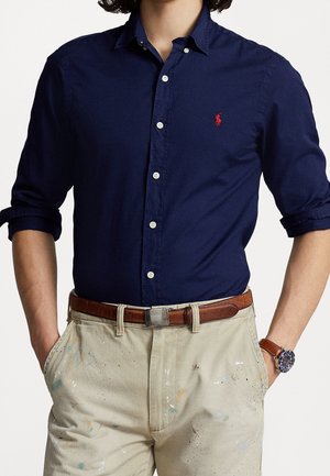 Man draagt een marineblauw overhemd met knopen en opgerolde mouwen, beige verfspattenbroek, bruine leren riem en polshorloge met bruine band.