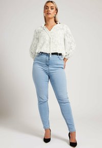 Blusa bianca con motivo floreale e colletto a cravatta, abbinata a jeans skinny a vita alta blu chiaro e tacchi neri a punta.