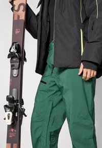 Slidinėjimo slidės su rudais viršutiniais elementais ir juodomis fiksavimo detalėmis, laikomos šalia žalių sniego kelnių. Striukė juoda su geltonais akcentais ir užtrauktukais.