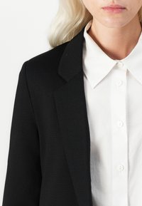 Svart blazer med en strukturert krage, kombinert med en hvit skjorte med knappestolpe. Glatt stofftekstur, med hvite knapper langs skjortefronten.