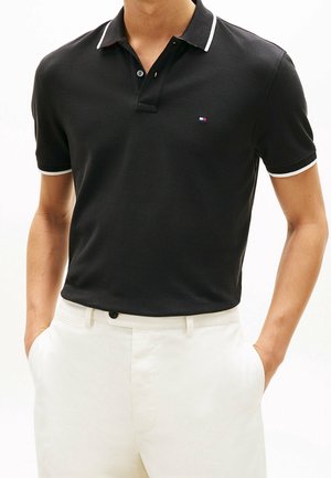 Homme portant un polo noir à manches courtes avec des bordures blanches et un pantalon blanc, les mains dans les poches.